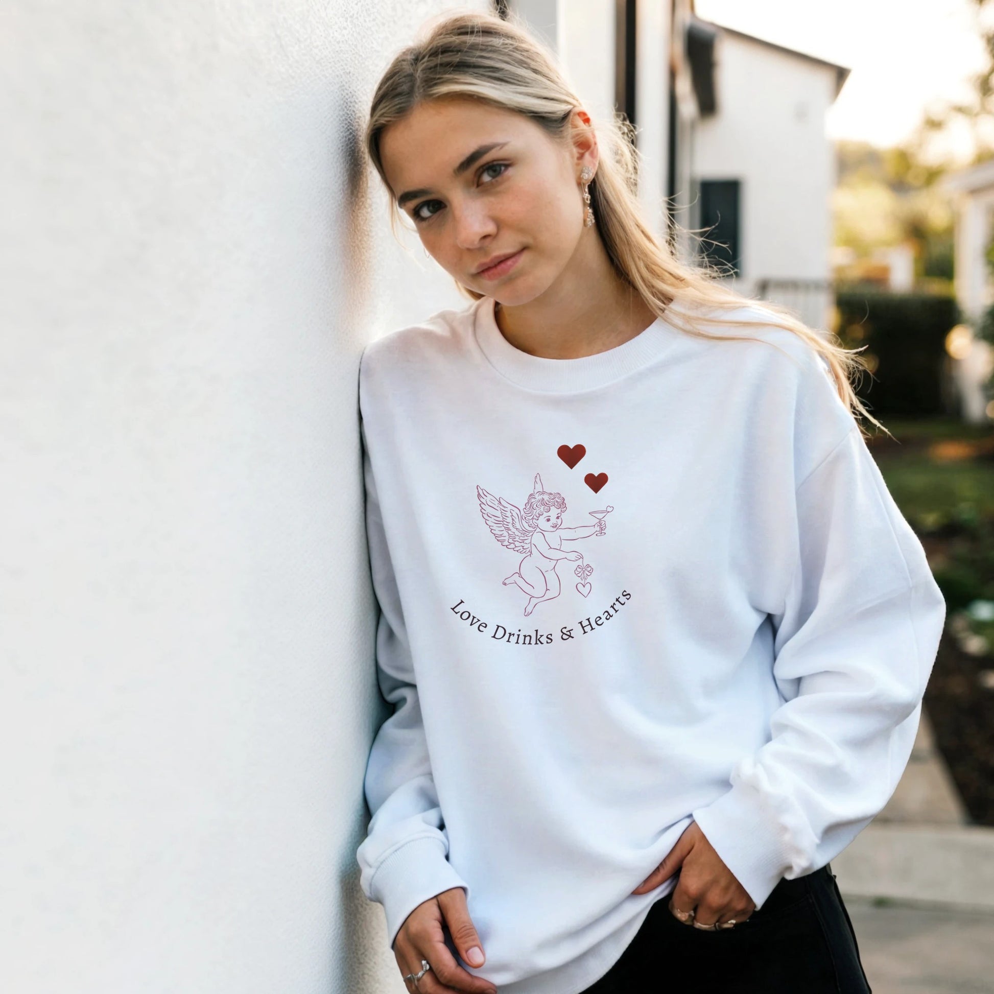 Woman wearing Love Sweatshirt with Love Drinks & Hearts design, standing against a wall outdoors.
