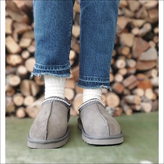 Person wearing Ugg Tasman II Slipper Mole with jeans and white socks, standing on a green surface in front of a wood pile.