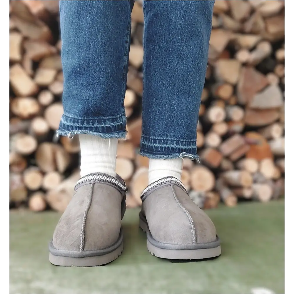 Person wearing Ugg Tasman II Slipper Mole with jeans and white socks, standing on a green surface in front of a wood pile.
